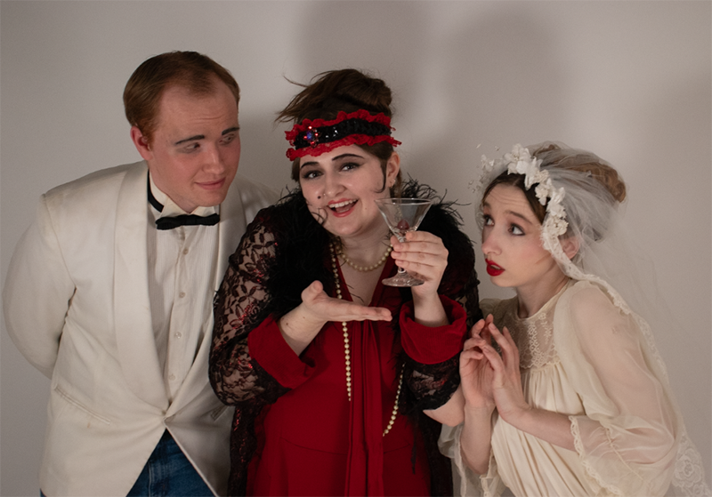 three people, all dressed in period costume, pose for a photo. man on left is wearing a white tux, woman in middle is wearing a red and black 1920s style costume and holding a a martini glass and woman on right is wearing a white dress.