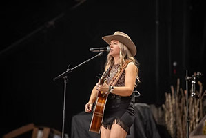 Woman with long blonde hair wearing a cowboy hat and playing a guitar