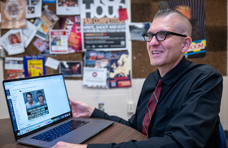a man with a mostly bald head and black glasses sits at a table and shows a laptop screen. He's wearing a black dress shirt and red striped tie