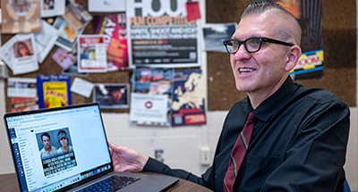 a man with a mostly bald head and black glasses sits at a table and shows a laptop screen. He's wearing a black dress shirt and red striped tie