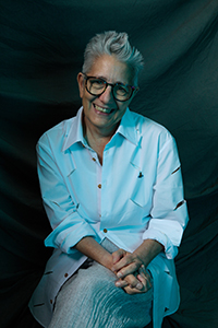 a photo of woman with short white hair, glasses and a white button up shirt. She's sitting with her legs crossed and hands folded on her lap
