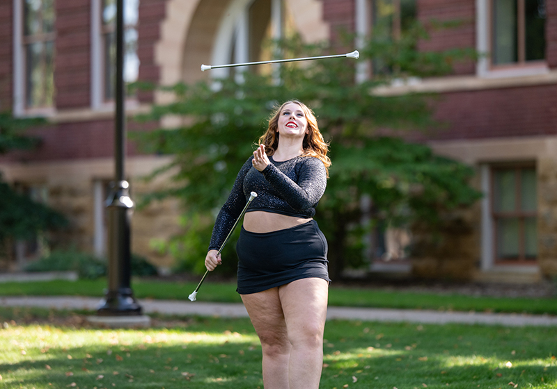 a female baton twirler stands outside a brick building. She's wearing a two piece black outfit and is tossing a baton in the air.