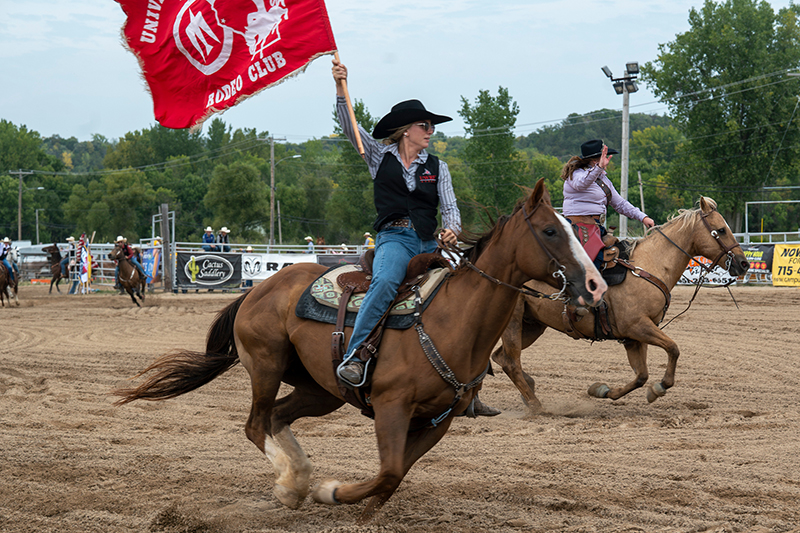 a woman rides a brown horse in a rodeo arena. She's wearing jeans, a black vest and black cowboy hat and is carrying a flag that says "UWRF Rodeo Club"