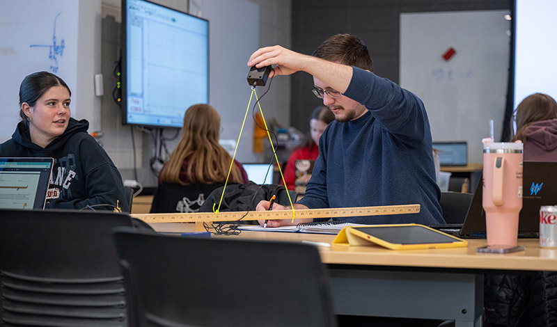 a male student uses a string measuring device as part of a math class while a female student sitting at the same table looks on