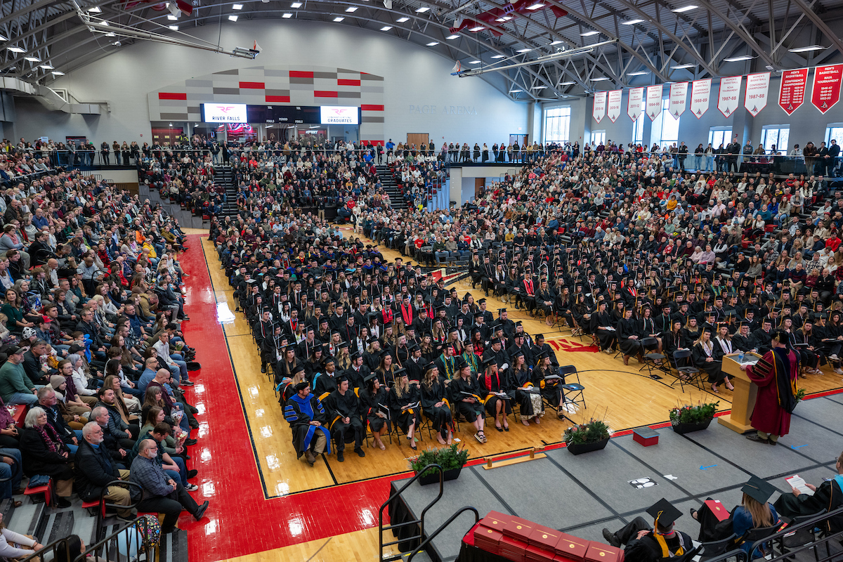 a photo of the crowd at Commencement. GRaduates are seated in chairs on the gym floor, while families and attendees fill the stands.