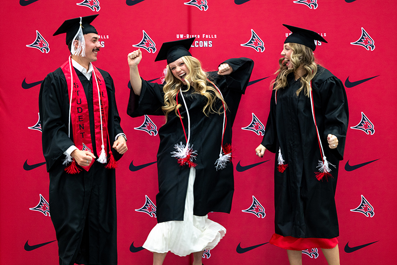 three students wearing black caps and gowns and various red and white cords pose in front of a backdrop that has the Nike logo and Falcon heads across it.