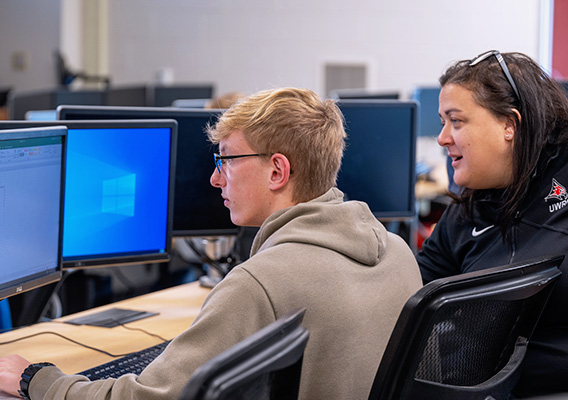 A male student sits at a computer with their professor behind them during class