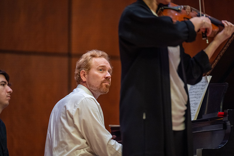 a man sits at a piano and plays while a person plays the violin in the foreground