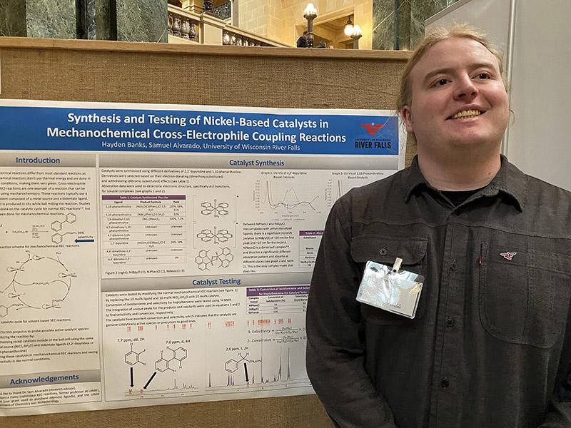 a person wearing a dark grey button up and nametag stands in front of a chemistry research poster
