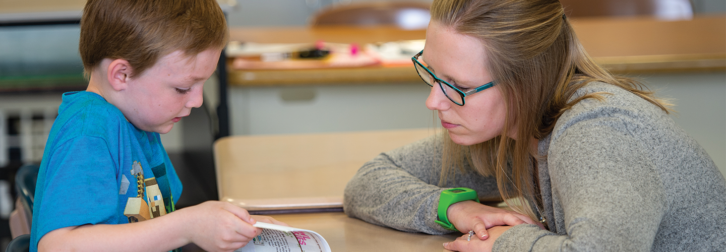 teacher helping a student read a book at their desk