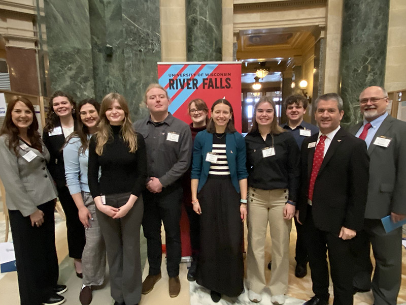 A group of people pose in business clothing in front of a sign that says UW-River Falls.
