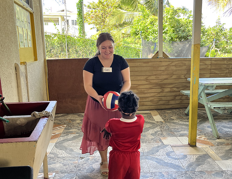 a woman wearing a black top, red skirt and nametag that says "Cori" faces the camera and holds a volleyball that she's handing to a young child wearing all red with their back to the camera
