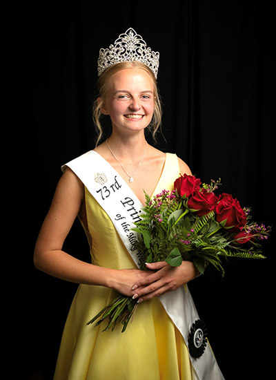 Smiling woman in a tiara and yellow gown wearing a “73rd Princess” sash and holding a large bouquet of red roses and greenery.