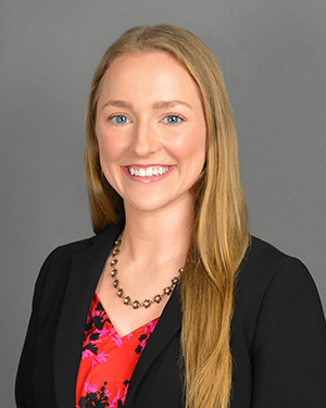 Jessica Moor. A woman with long blonde hair wearing a pink and red blouse with a black blazer in front of a grey background