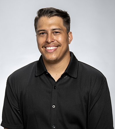 Jordan Kellogg headshot. A man with dark hair and facial hair is wearing a black button up shirt in front of a grey backdrop
