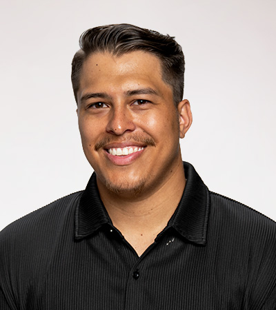 Jordan Kellogg headshot. A man with dark hair and facial hair is wearing a black button up shirt in front of a grey backdrop