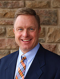 headshot of Kevin Westhuis, a white man wearing a blue blazer and orange and blue plaid tie standing in front of a brick wall