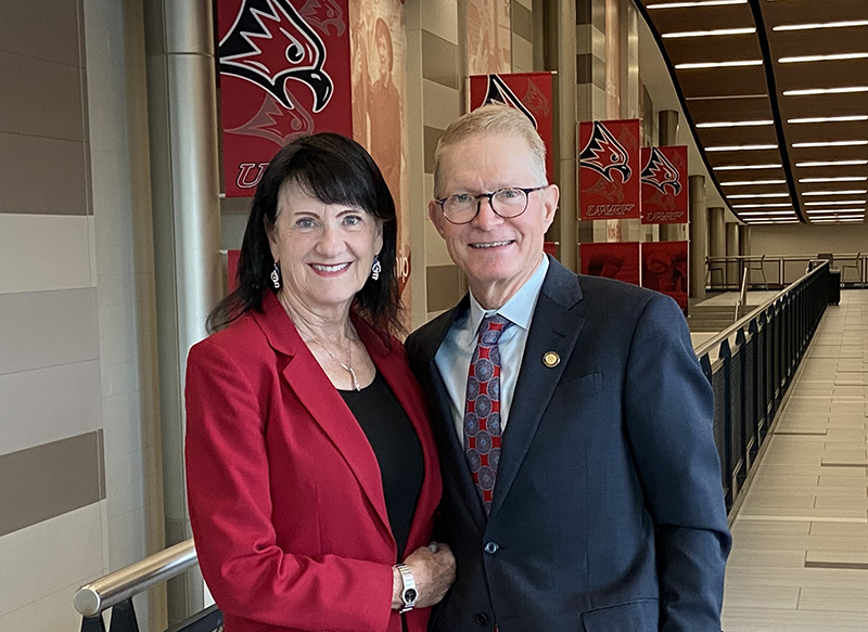 Karen and Paul Dykstra. Two people pose together, a white woman with long black hair wearing a red blazer and black shirt and a white male with blonde hair wearing a navy blue sportcoat.