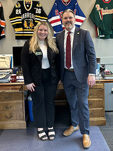 two people stand in an office with numerous hockey jerseys hanging on the wall, one female wearing a black pant suit and one male wearing a grey suit and red tie