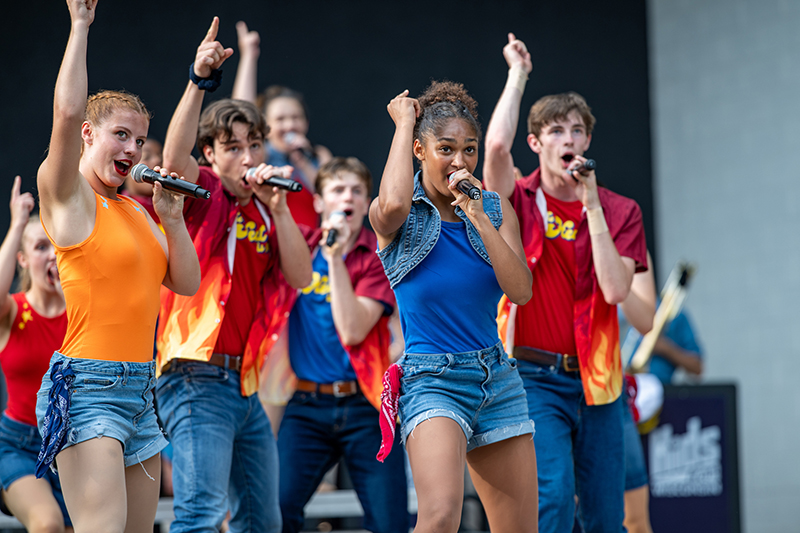 a group of teenagers perform on stage. They're holding microphones and signing and wearing brightly colored tops and denim shorts