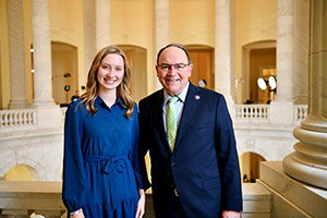 two people pose together in a government building, one female in a blue dress and one male in a navy suit and light green tie