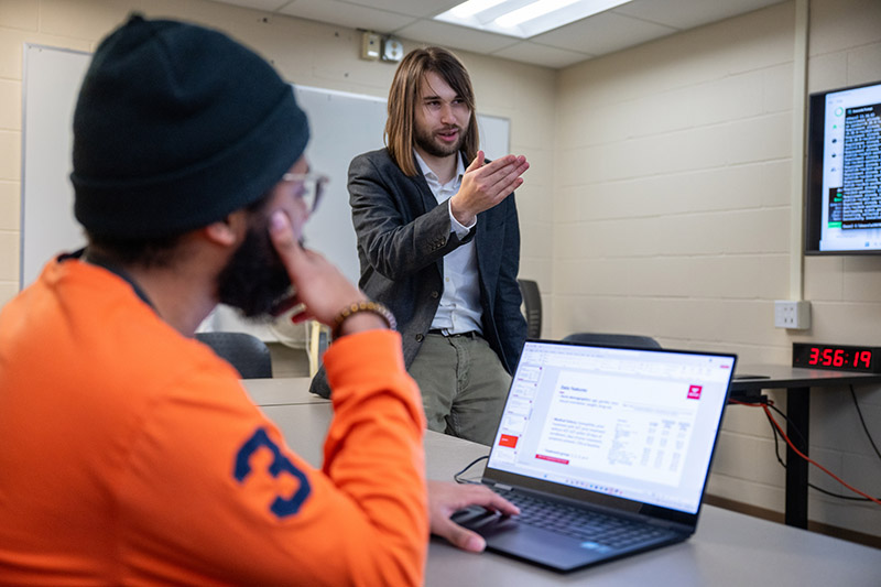 a faculty member wearing a grey sport coat points at a board while a student wearing an orange shirt and black hat listens while sitting in front of a laptop