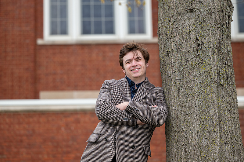 a man wearing a grey suit coat leans against a tree
