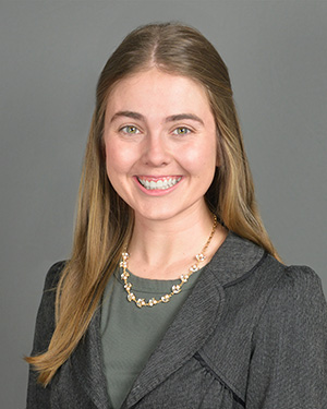 Michelle Stangler. A woman with long brown hair wearing a green blouse and gray blazer in front of a gray background