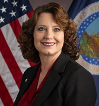 A portrait of a woman with dark brown shoulder length curled hair. She's wearing a red blouse and a black blazer and is seated in front of the United States Flag and a flag with a U.S. seal on it. 