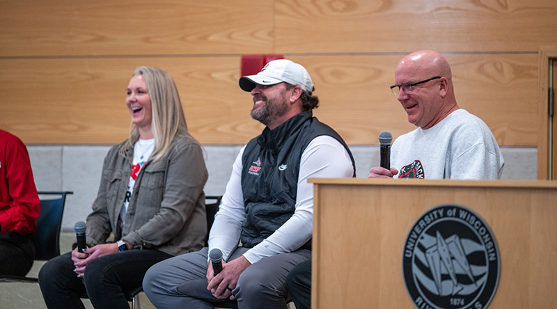 three people sit on a stage, a blond woman to the left, with a man wearing a white hat in the middle and a man wearing a white sweatshirt sits on the right. all are holding microphone.