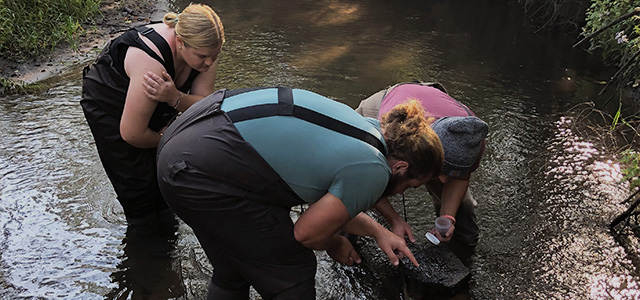 Three students stand in the Kinnickinnic River analyzing sand at the bottom of the river