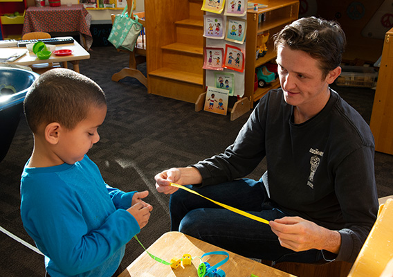 A student teacher holds out a measuring tape for a preschooler while in a classroom