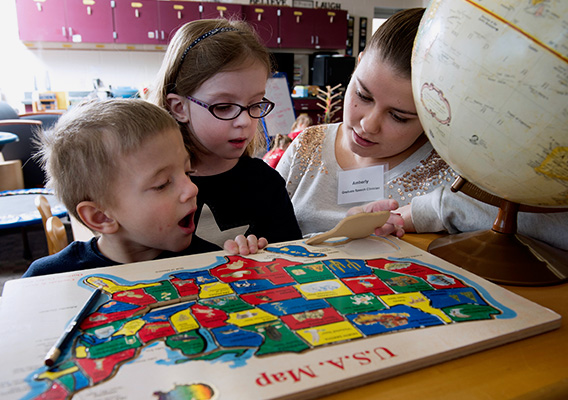 Two preschoolers look at a map of the USA while a student teacher watches