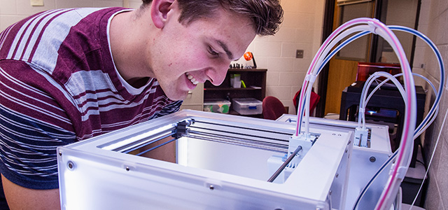 A male student looks down into a 3D printer to view the progress