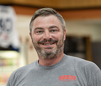 headshot of Pat Ptacek. A man with short brown and grey hair and brown and gray facial hair. He's wearing a grey t-shirt that says "Ptacek's IGA" in red text.