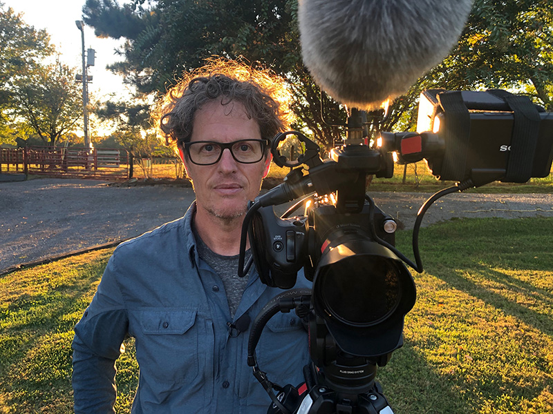 a man with curly grey hair poses for a photo while standing behind a film camera and microphone