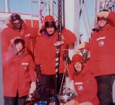 A group of people are at the South Pole. They are standing with scientific equipment and all wearing red and black snowgear.