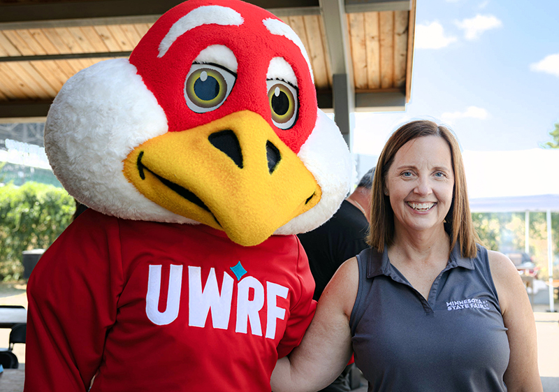 a woman with shoulder length brown hair and wearing a sleeveless grey shirt, stands next to UWRF's Freddy Falcon mascot