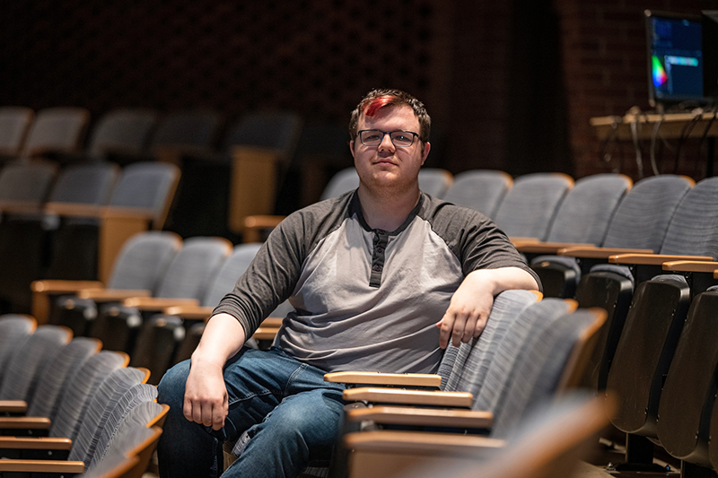 A college aged man sits in a chair in a theatre.