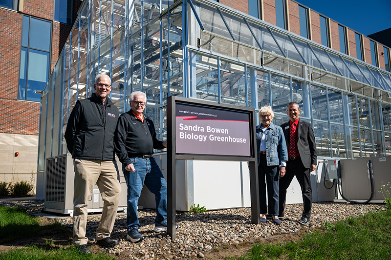 Four people pose with a sign in front of a greenhouse. The sign says "Sandra Bowen Biology Greenhouse."