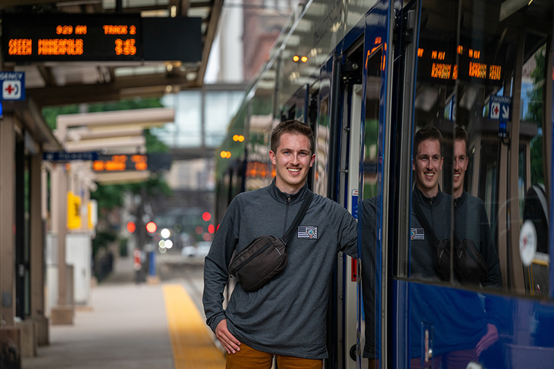 a man wearing a grey sweatshirt and a black shoulder bag leans against a metro train