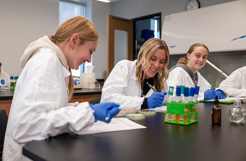 three students wear white lab coats and blue medical gloves while working in a chemistry lab. A green test tube holder sits on the lab table, along with Petri dishes, as they write on paper. 
