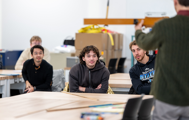 three male students sit at a table while listening to a professor talk