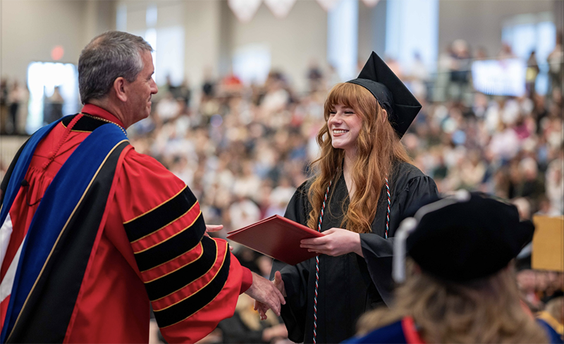 a female student wearing Commencement regalia is handed a diploma by the Chancellor, who is wearing a red and black robe