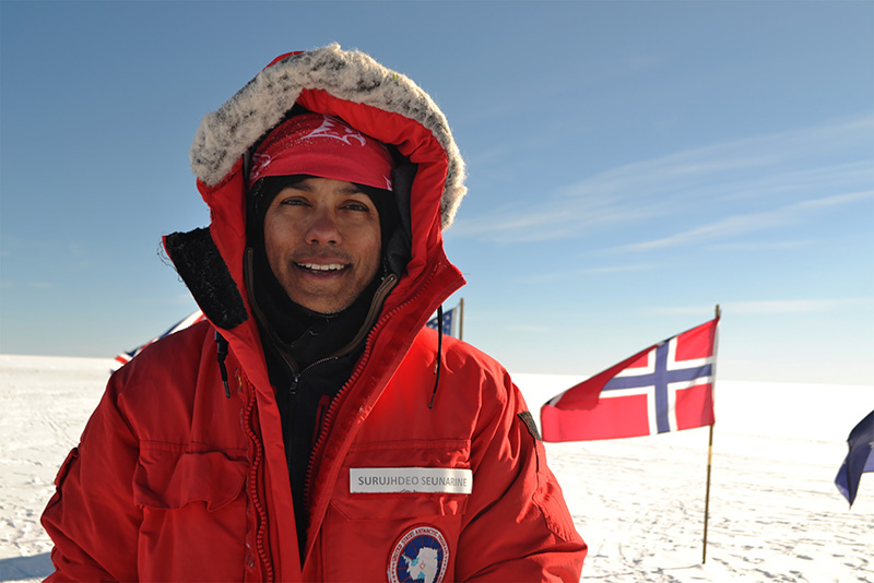 a man stands at the south pole with a flag in the background. He's wearing red snowgear.