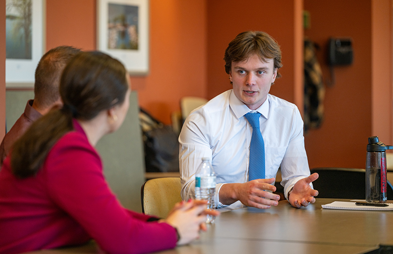 a white male wearing a white dress shirt and blue tie sits at a table and discusses his plans with two other people