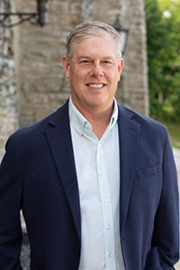 Steven Hahn headshot. A white man with short grey hair poses for a headshot outdoors and is wearing a navy blue blazer and light blue dress shirt.