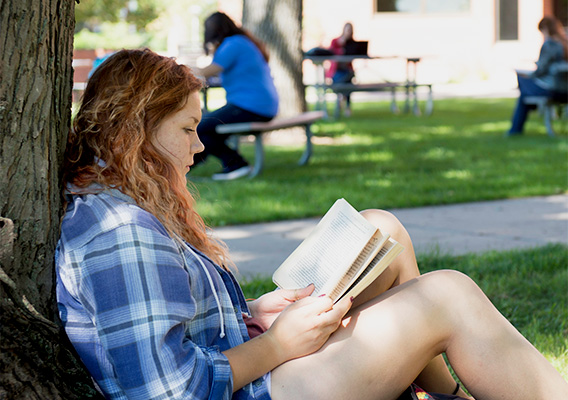 A student sitting under a tree reading a book on campus