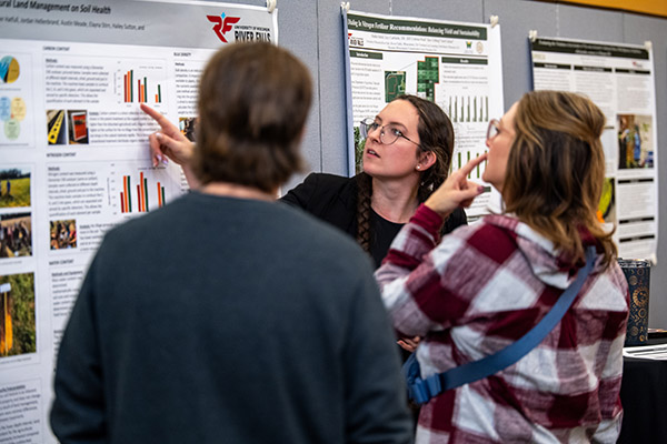A student points to a posterboard with a research presentation on it while two people look on