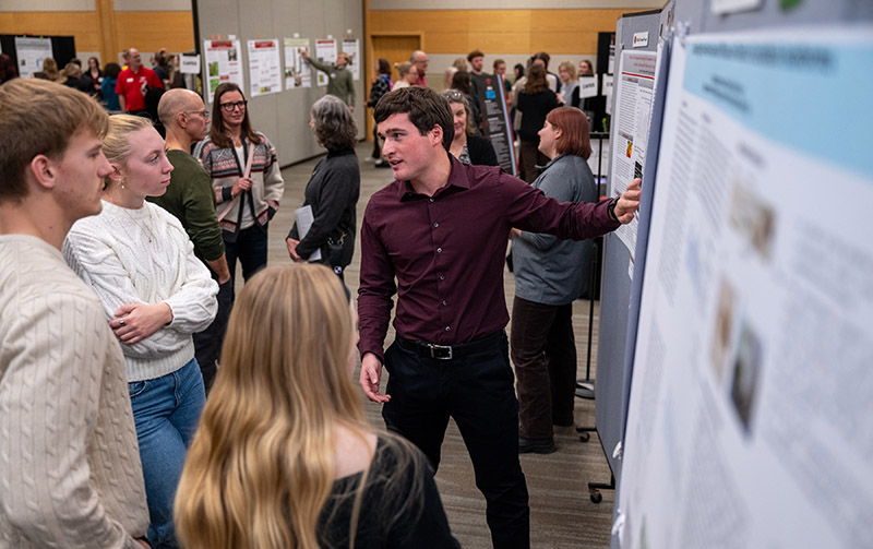 a white male college student stands in front of a poster displaying research results and explains his work to three onlooking students